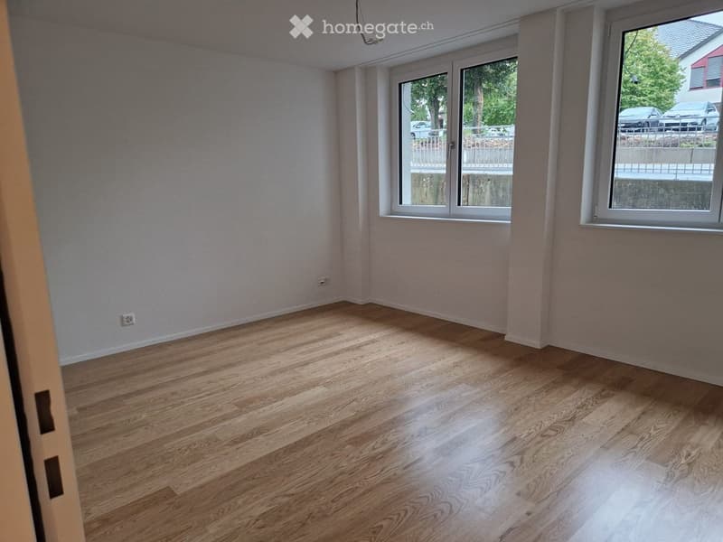 Empty room with wooden flooring and large windows, showing natural light and a view of the outside area.