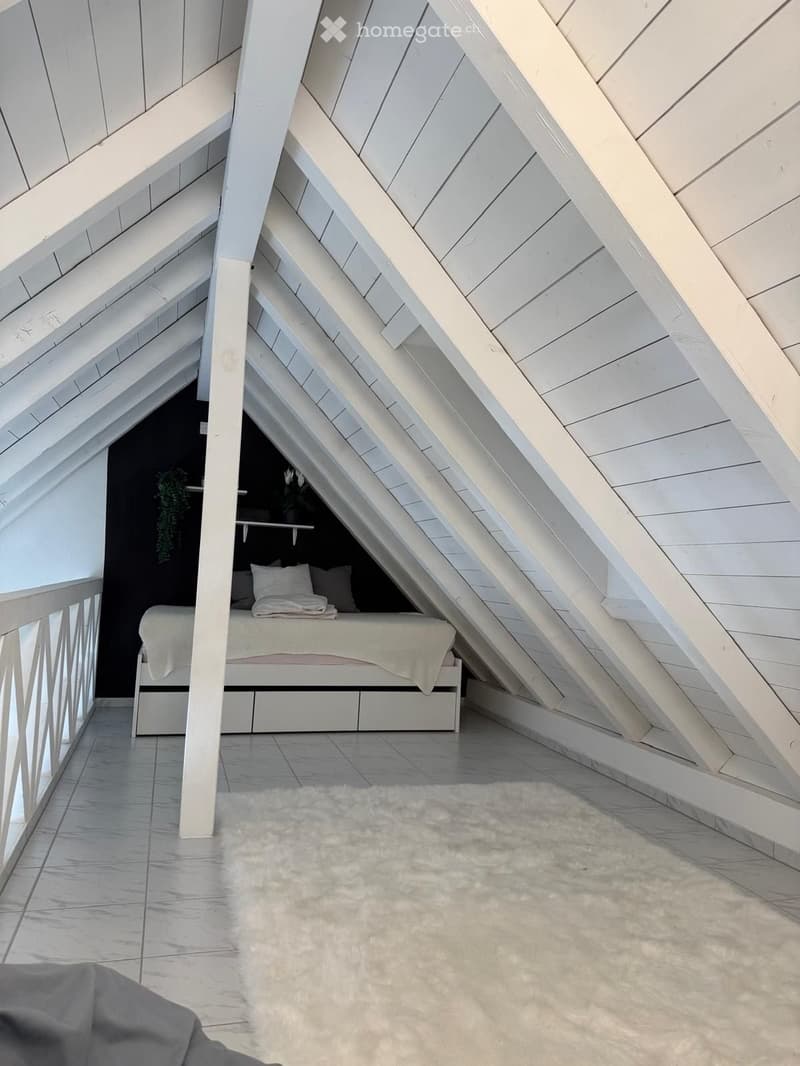 Attic bedroom with slanted wooden ceilings, white bed with pillows and blanket, white rug, and built-in shelves with decorative items.