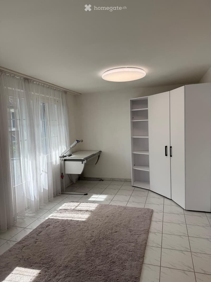 A well-lit room with a tiled floor, a large window with white curtains, a round ceiling light, a white desk with a lamp, a tall white cabinet with shelves, and a brown shag rug.