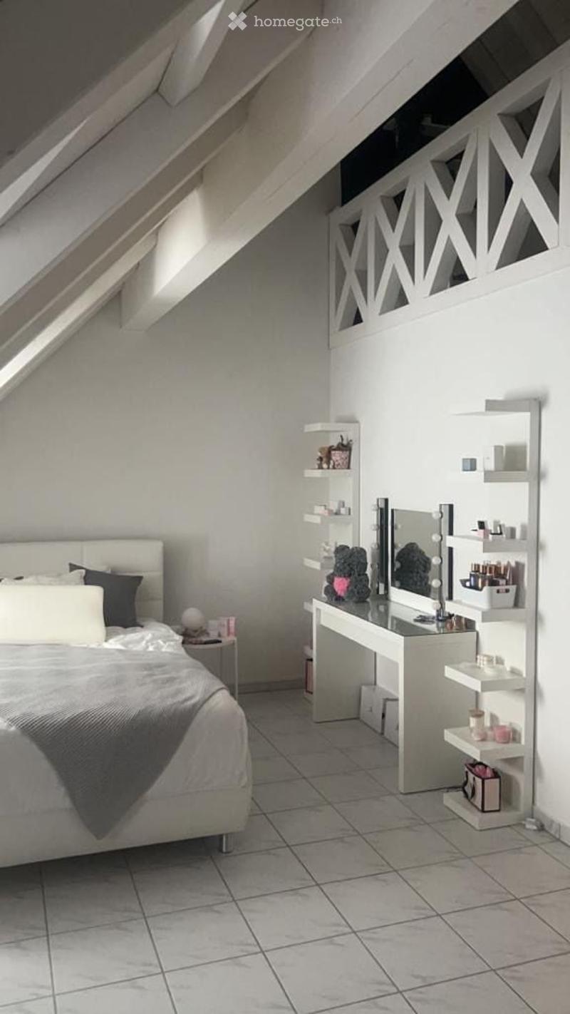Modern attic bedroom with a white bed, grey blanket, vanity with mirror, shelves, and nightstand. The room features exposed wooden beams and skylights.