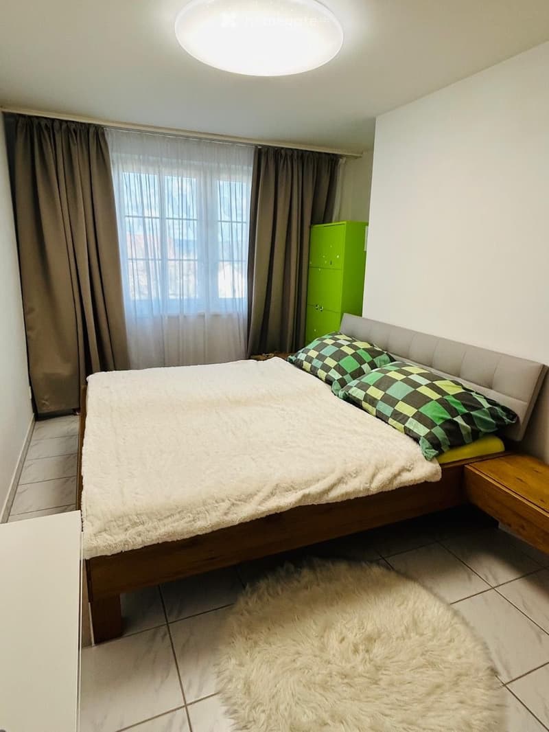 A cozy bedroom with a large bed, fluffy white bedding, patterned pillows, a wooden nightstand, and a green cabinet. The room features tiled flooring, a fluffy white rug, and large windows with grey curtains.