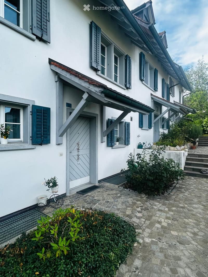 Two-story white house with grey shutters, stone pathway, small garden, bicycle, and potted plants near the entrance.
