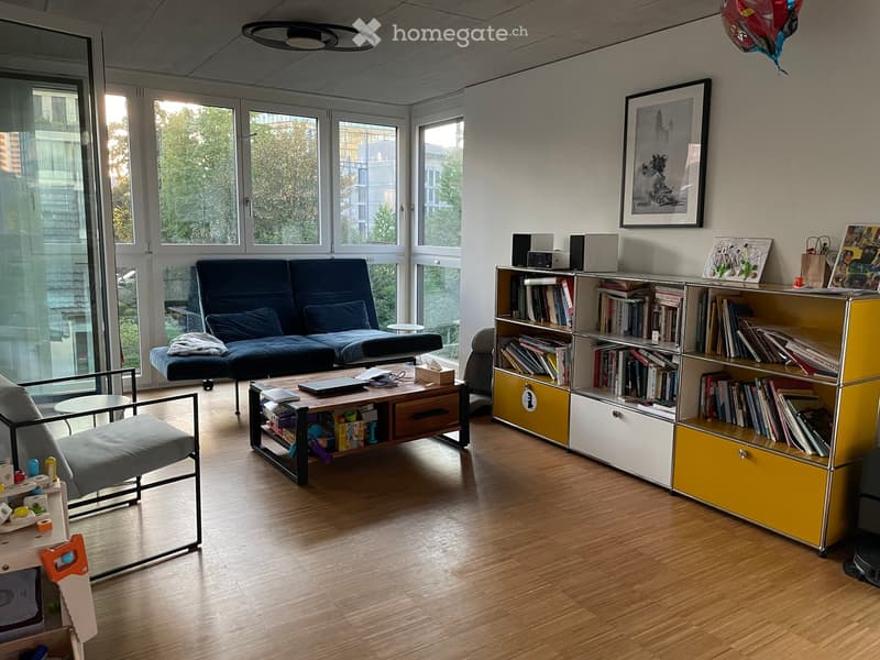 Modern living room with wooden flooring, large windows, a blue sofa, a coffee table, a bookshelf, and a chair.