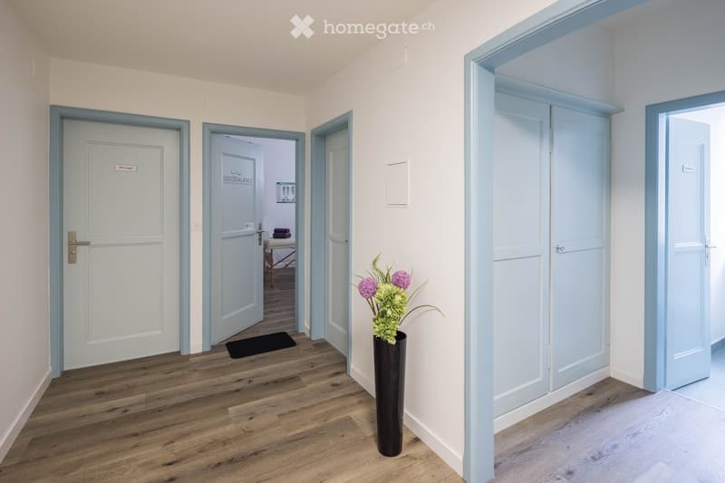 A clean, well-lit hallway with light blue doors and a wooden floor. A black vase with flowers is placed near the center of the hallway.