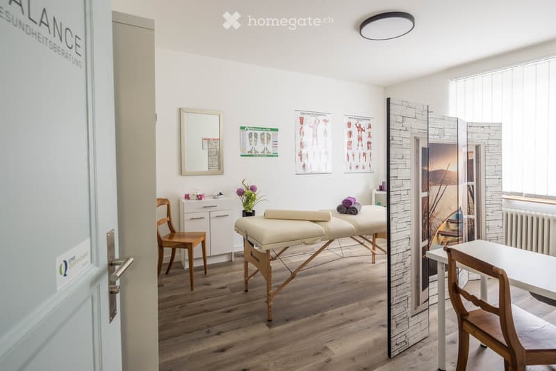 A well-lit room with wooden flooring, two massage tables, a wooden chair, a small cabinet, a mirror, and a table with a flower pot.