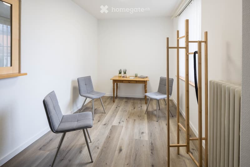 A small, well-lit room with light wooden flooring, white walls, and a large window. It features a small wooden table with two matching chairs, a coat rack, and a radiator.