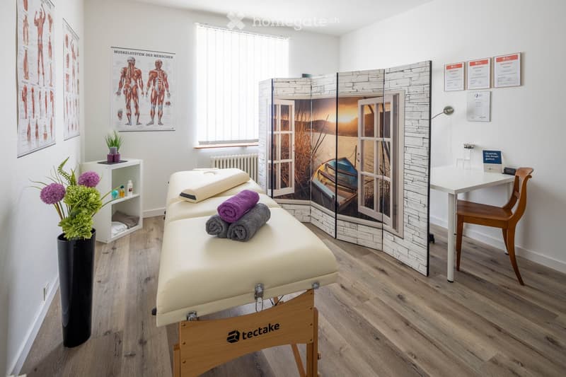 Well-lit room with wooden flooring, two massage tables, a small table with a chair, a vase with flowers, a screen, and posters on the wall.