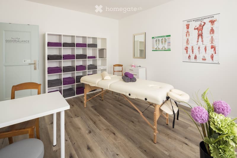 A clean, well-lit massage room with wooden flooring, two massage tables, a shelf with neatly stacked towels, a small table, and a mirror on the wall.