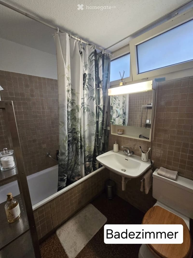 Bathroom with a bathtub, sink, toilet, mirror, and a shower curtain. The room features tiled walls and a wooden toilet seat.