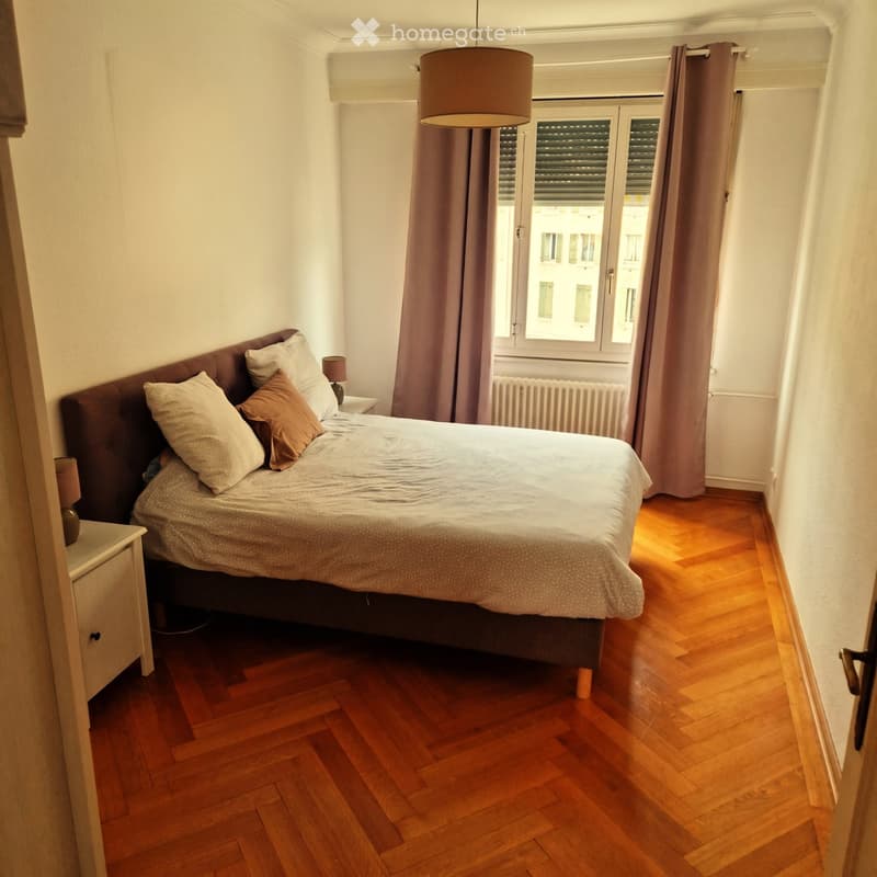 Cozy bedroom with wooden flooring, a large bed with pillows and a duvet, nightstands with lamps, and large windows with curtains.