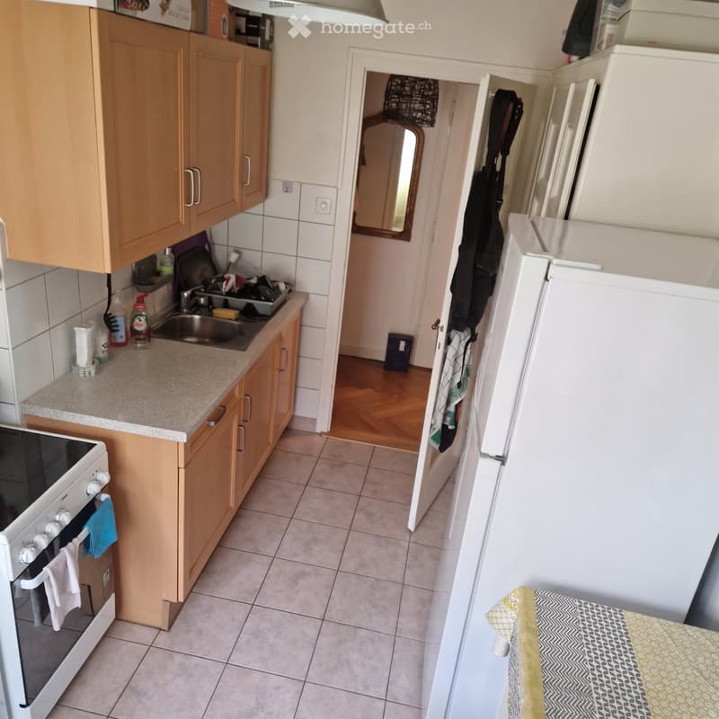 Compact kitchen with wooden cabinets, a sink, oven, refrigerator, and a small dish rack. The space features tiled floors and a countertop with various items.