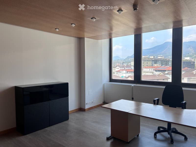 Modern office space with a large window offering a scenic view of the city and mountains. Features a wooden desk, office chair, and a sleek black cabinet.