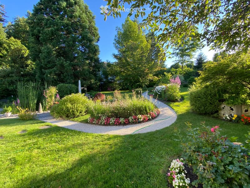 Well-maintained garden with a winding gravel path, lush green grass, colorful flowers, shrubs, and tall trees providing shade.