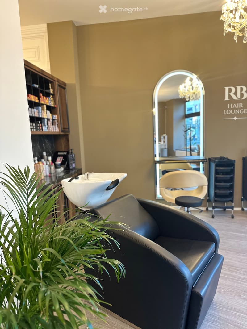 Modern hair lounge with a black leather sofa, white sink, beige chair, large arched mirror, wooden cabinet, and decorative plant.