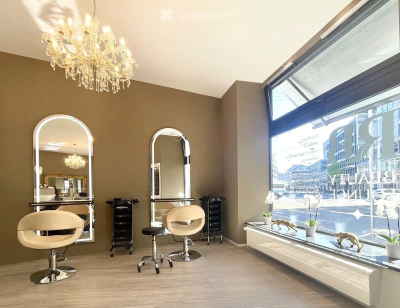 Modern salon with large windows, two stylist chairs, mirrors, a chandelier, and decorative flower pots on a console table.
