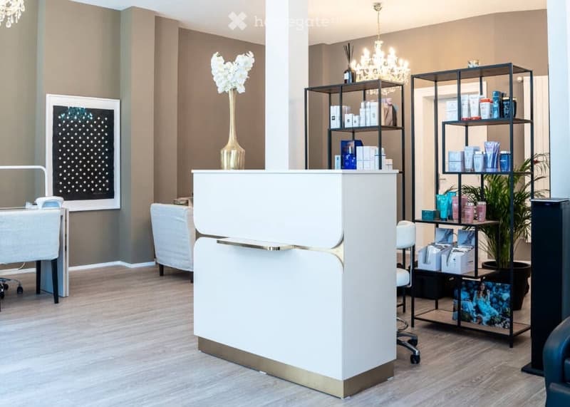Modern reception area with a white reception desk, a white chair, shelves with various products, a vase with white flowers, a chandelier, and a potted plant.
