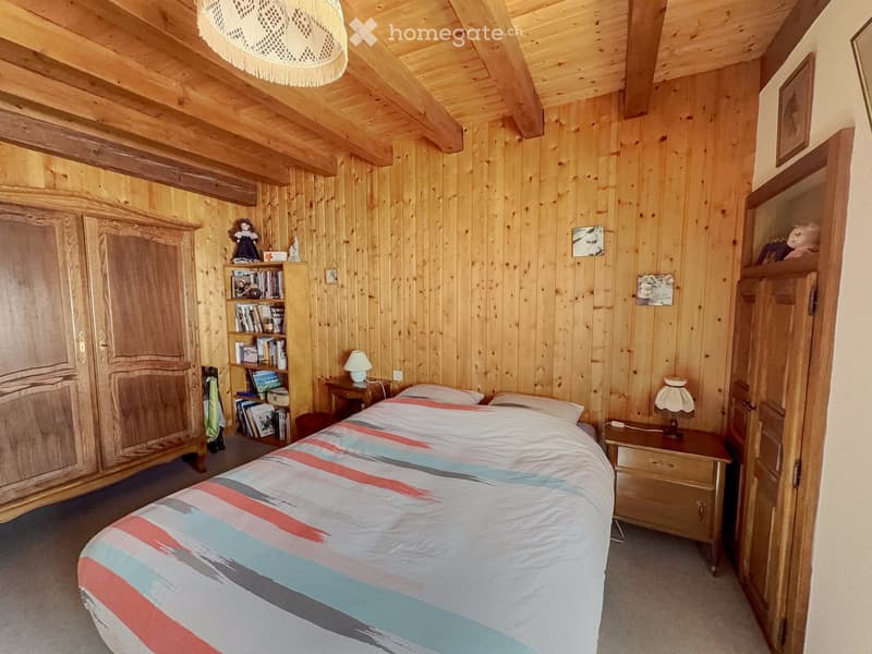 Cozy wooden bedroom with a bed, nightstands, lamps, bookshelf, and wardrobe. The room features exposed wooden beams and a warm, inviting atmosphere.