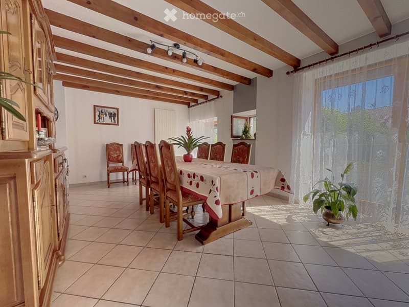 Dining room with wooden beams, tiled floor, dining table with chairs, cabinet, and plants.