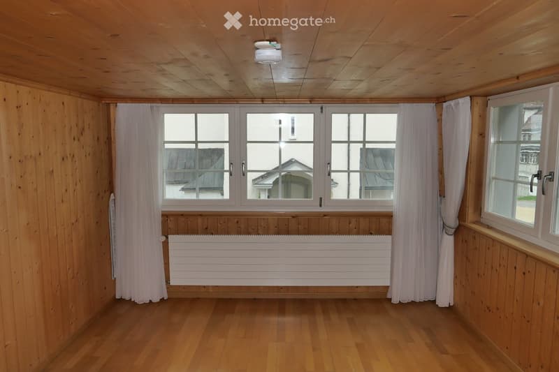 Empty room with wooden walls, wooden floor, large windows with white curtains, and a white radiator below the windows.