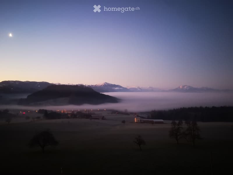 A serene landscape view at dawn with misty fields, distant mountains, and a small house in the foreground.