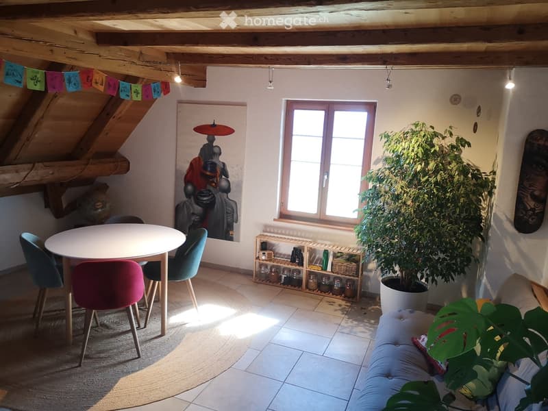 Cozy room with exposed wooden beams, a round white table with colorful chairs, a large potted plant, a shelf with jars, and a decorative sculpture on the wall.