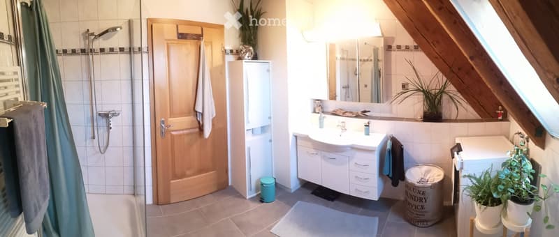 Bright bathroom with a bathtub, shower, sink, and mirror. Includes a wooden door, white cabinets, towels, and potted plants.