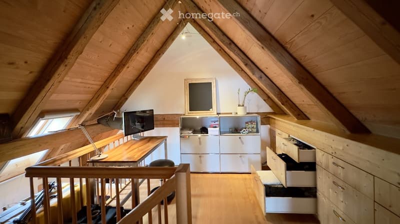 Cozy attic room with wooden beams, a desk with a screen and lamp, a cabinet with storage, and a small plant on top.