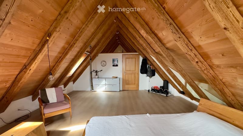 Bright attic room with wooden beams and slanted ceilings, featuring a bed, sofa, chair, small cabinet, clock, and exercise weights in the corner.