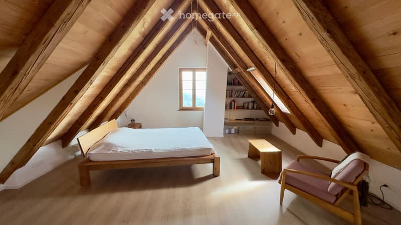 Cozy attic bedroom with wooden beams, slanted ceilings, a bed, a chair, a small table, and a built-in bookshelf.