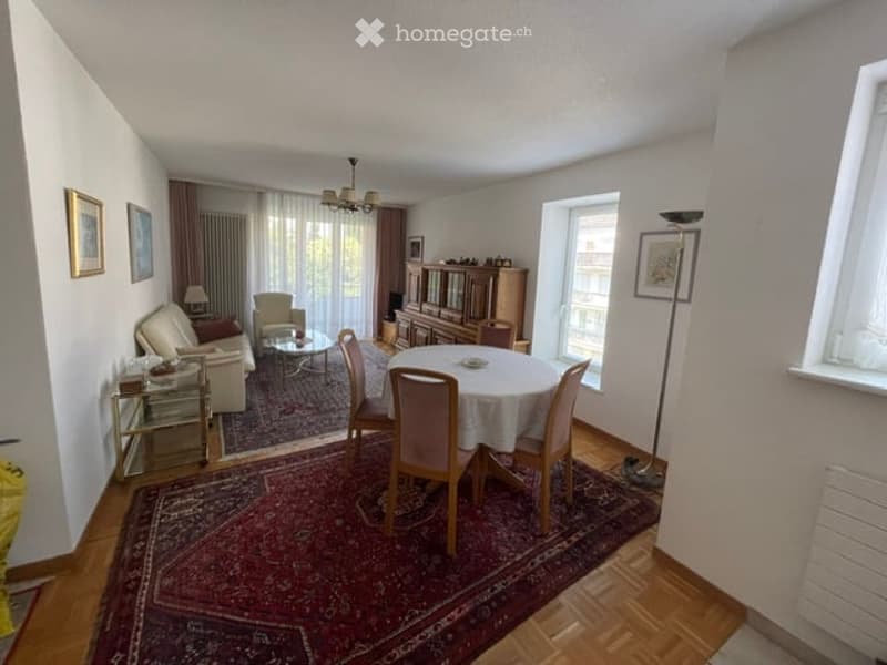 A well-lit living and dining area with a sofa, coffee table, dining table with chairs, and a floor lamp. The space features wooden flooring, large windows with curtains, and decorative picture frames on the walls.