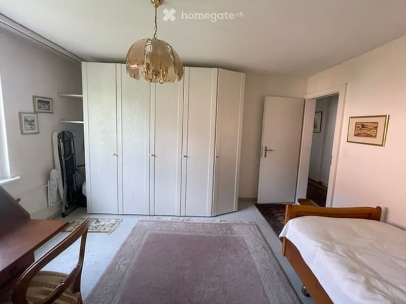 Bright bedroom with white walls, a large built-in wardrobe, a wooden bed, a chair, a small table, and framed pictures on the wall.