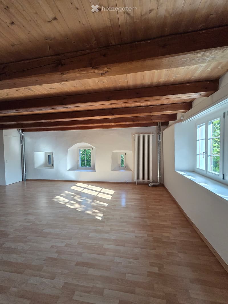 Empty room with wooden floor and exposed wooden ceiling beams. Features include white walls, multiple windows, radiator, and a small arched window.