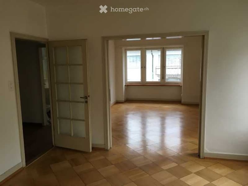 Empty room with wooden flooring, large windows, and a door leading to another space.