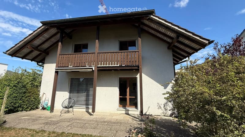 Two-story house with a balcony, wooden railing, and a covered entrance area. The house has a modern design with large windows and a sloped roof.