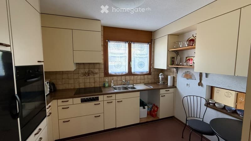 A small, well-lit kitchen with cream-colored cabinets, a dark countertop, and a window with floral curtains. The kitchen features a sink, a refrigerator, an oven, and a small dining area with a chair and table.