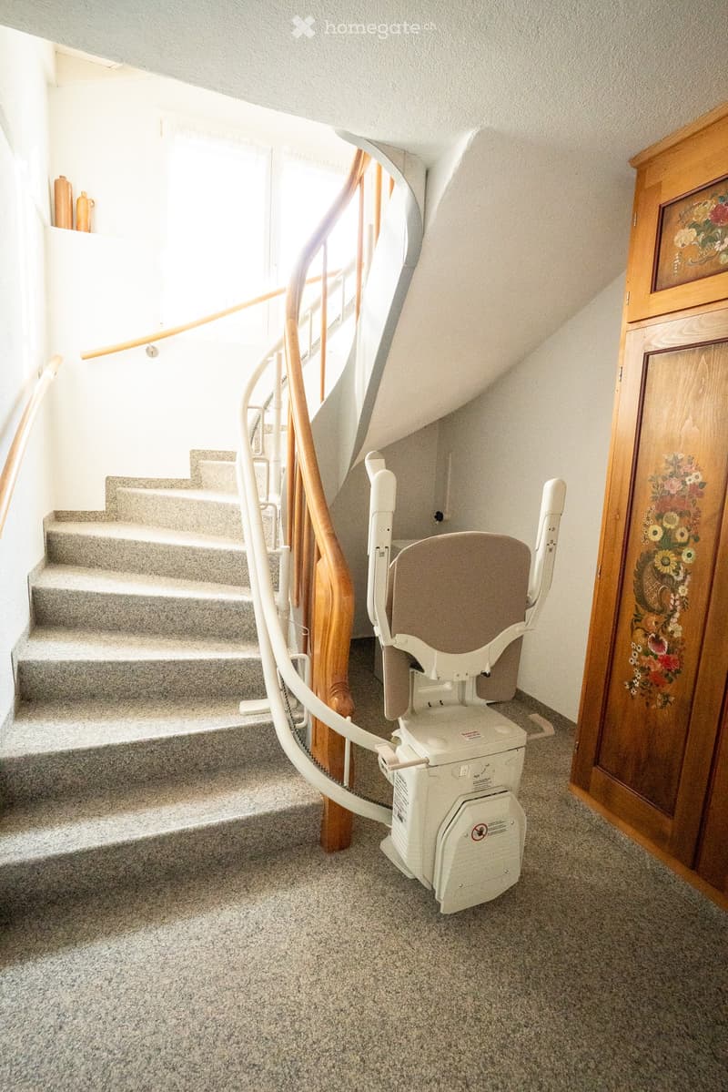 Carpeted hallway with a staircase, wooden handrail, and a stair lift installed on the stairs.