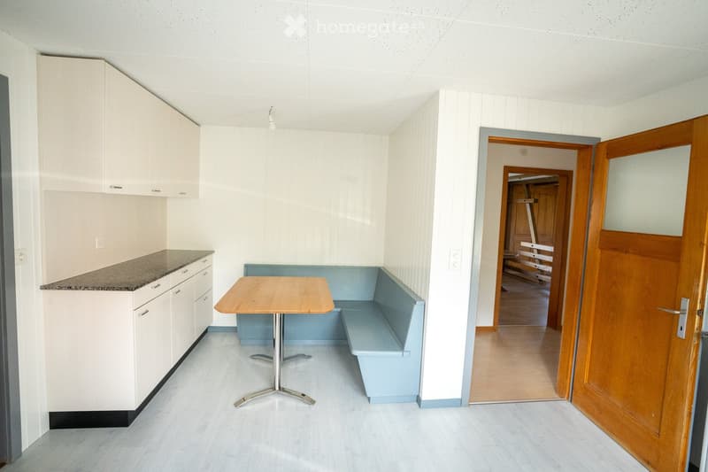 Bright, modern room with white cabinets, a countertop, a small round table with a bench seating area, and a wooden door leading to another space.
