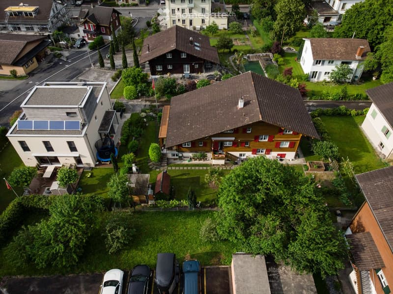 Aerial view of a residential neighborhood with multiple houses, gardens, and parked cars. Houses feature varied architectural styles, including modern and traditional designs, surrounded by greenery and well-maintained lawns.