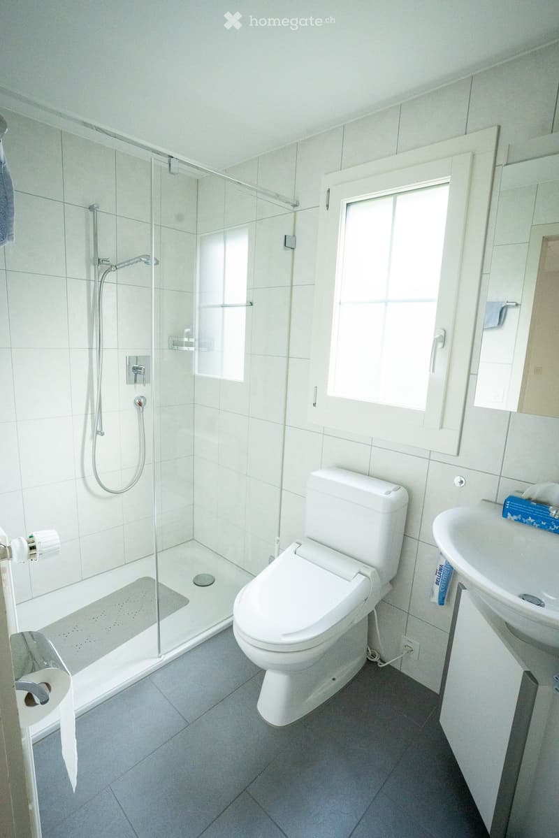 Modern bathroom with white tiles, a toilet, a sink, a glass-enclosed shower, and a mirror above the sink.