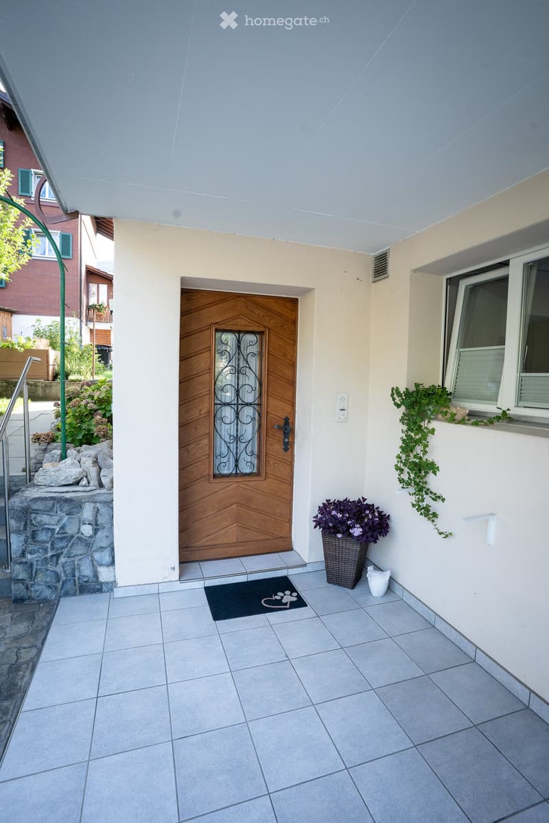Entrance area with tiled floor, wooden door with glass panel, doormat, potted plant, and window with climbing plant.