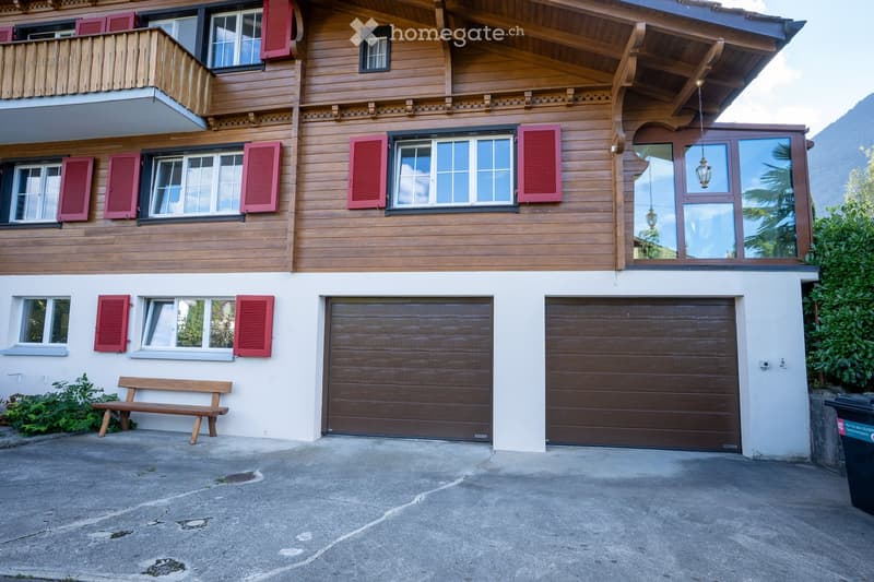 A two-story wooden house with red shutters, large windows, and two garage doors. The house features a balcony on the upper floor and a small wooden bench near the entrance.