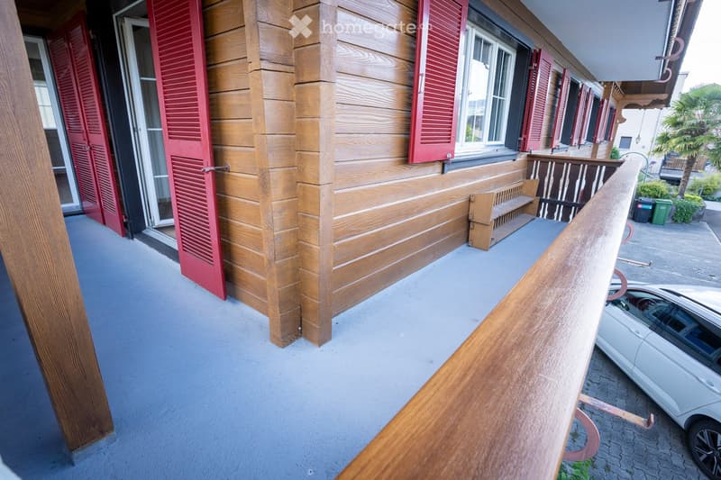 Balcony with wooden railings, shutters, and a bench, overlooking a parking area with a car.