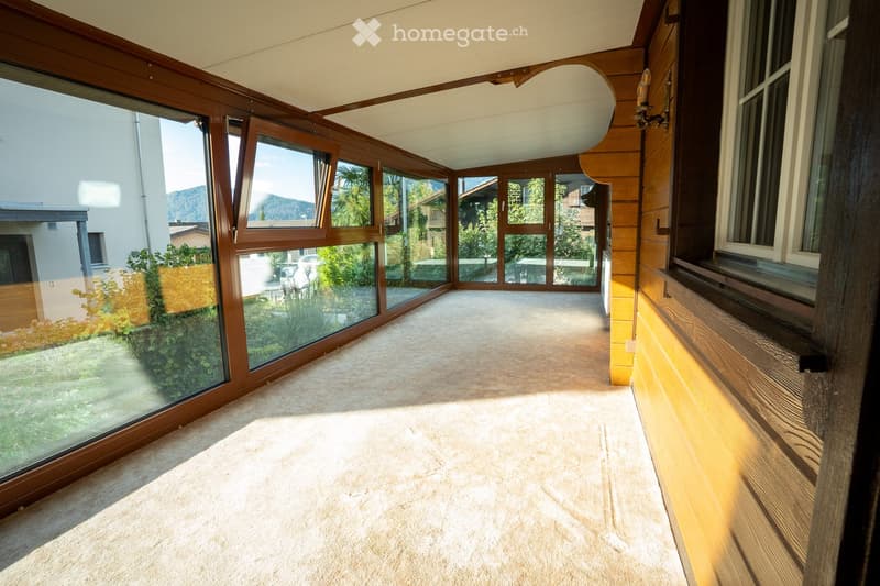 Bright hallway with large glass windows and wooden paneling, leading to an outdoor area with a view of greenery and mountains.