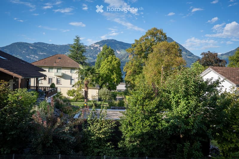 A lush garden with various plants and trees, featuring a view of a house with a balcony and surrounding mountains.