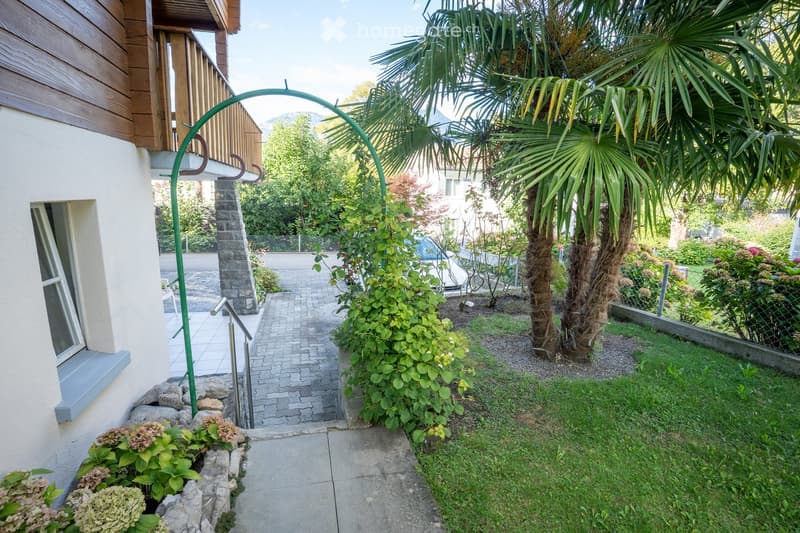 Garden pathway lined with palm trees and greenery, leading to a house entrance with a staircase and railing.