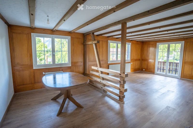 Bright, wooden-paneled room with exposed wooden beams, large windows, and a round wooden table with a bench.