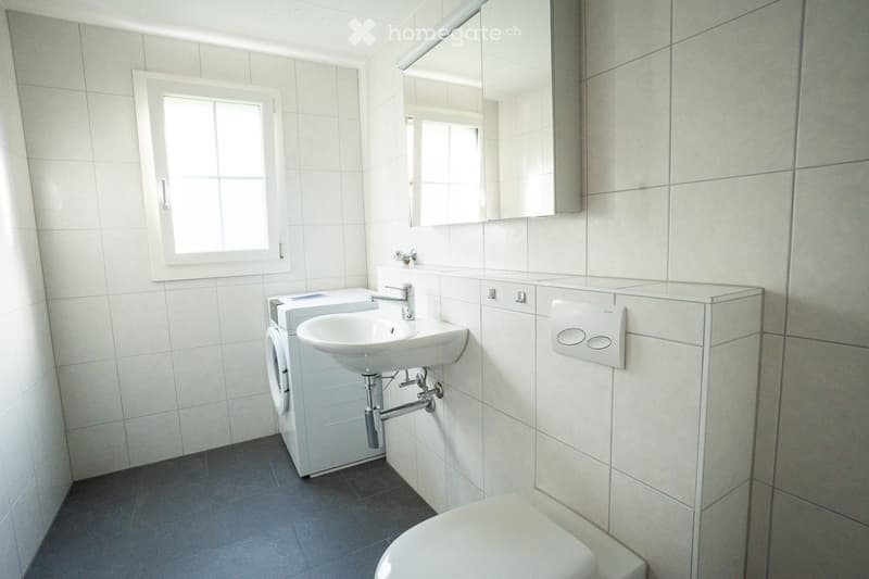 Modern bathroom with white tiles, a sink, toilet, mirror, and a washing machine in the corner.