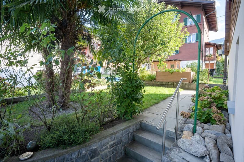 Garden area with steps, greenery, and a curved metal archway leading to a pathway.
