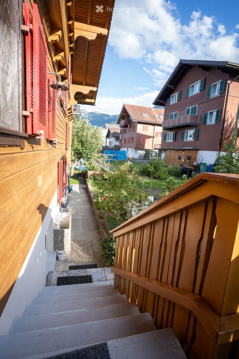 A narrow, paved pathway lined with wooden railings and greenery, leading to a staircase and surrounded by traditional wooden houses with balconies and shutters.