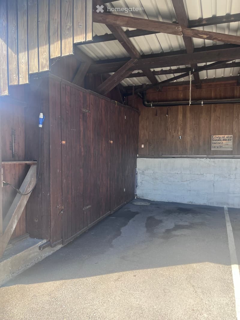 Empty stable with wooden stalls, concrete floor, exposed wooden beams, and a sign on the wall.